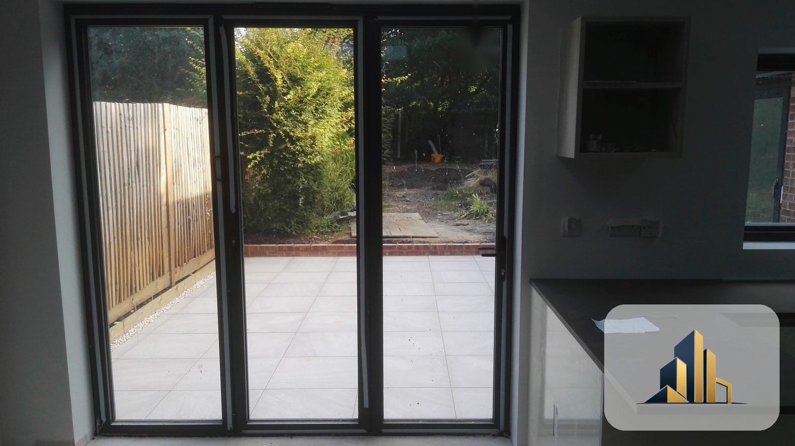 Finished open-plan kitchen and living space looking out through bifold doors to the tiled terrace, Raynes Park